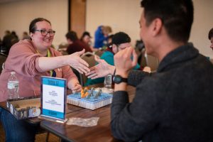 The Social Benefits of Board Gaming: Two players shaking hands across a table during a friendly board game tournament at the Great Plains Game Festival. A festive and welcoming atmosphere with participants engaged in various games in the background.