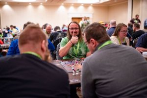 The Social Benefits of Board Gaming: A smiling player giving a thumbs-up while playing a board game at the Great Plains Game Festival. The room is filled with attendees enjoying their games, showcasing the lively and inclusive event environment.