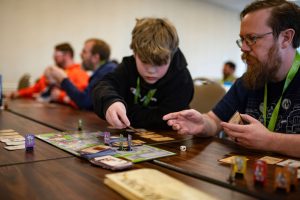 A young boy and an adult mentor focus intently on a board game at the Great Plains Game Festival. The mentor offers guidance while the child makes a strategic move, highlighting the intergenerational connection and learning that board gaming fosters.