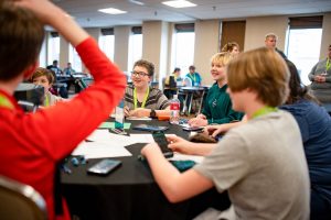 A young boy and an adult mentor focus intently on a board game at the Great Plains Game Festival. The mentor offers guidance while the child makes a strategic move, highlighting the intergenerational connection and learning that board gaming fosters.