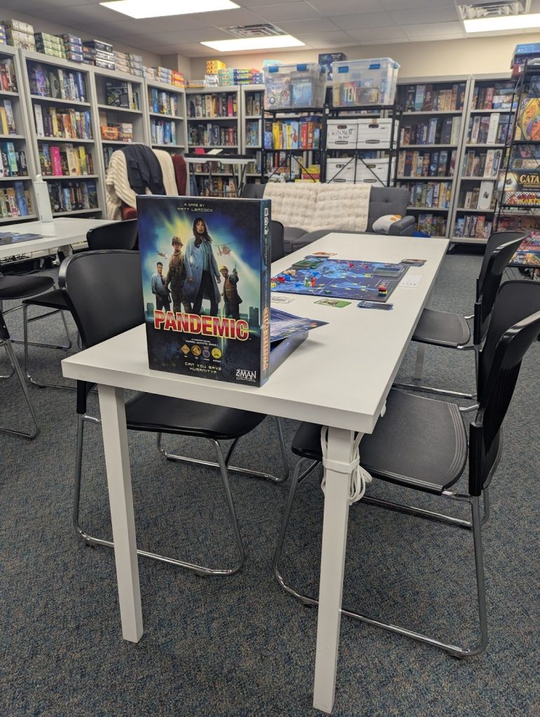 Pandemic set up for a community game night on a table for demo surrounded by shelves lining the walls filled with board at the GPG HQ.